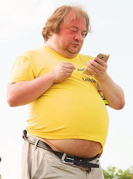 An overweight man wih his belly peaking out under his shirt as he looks at his phone.